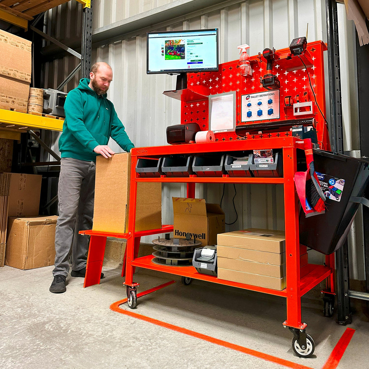 A man standing next to a red multi-tiered packing bench with a workstation setup, including a computer monitor, in a warehouse environment.